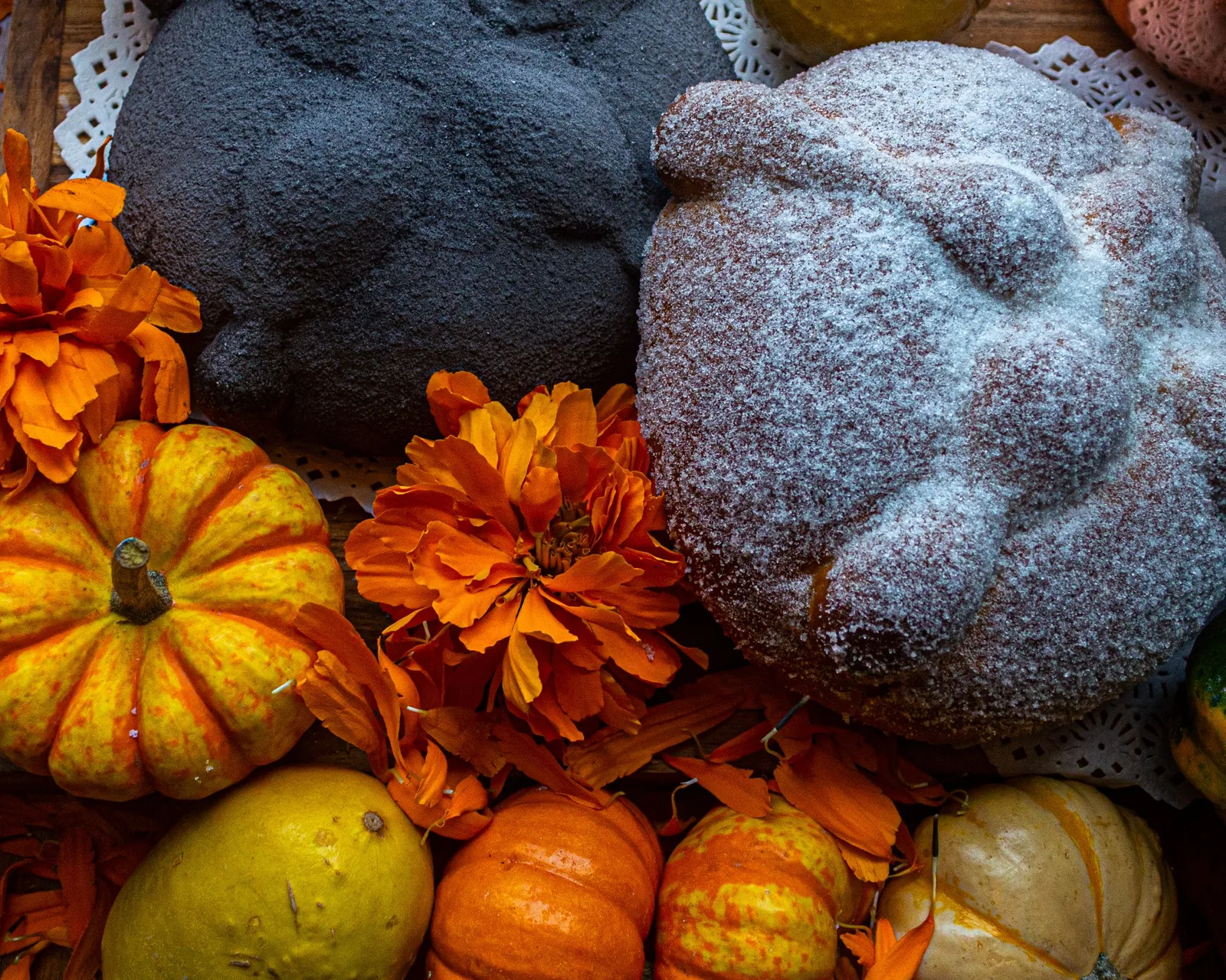 Un pan de muerto espolvoreado de azúcar junto a un pan de muerto negro, con cempasúchil y calabazas